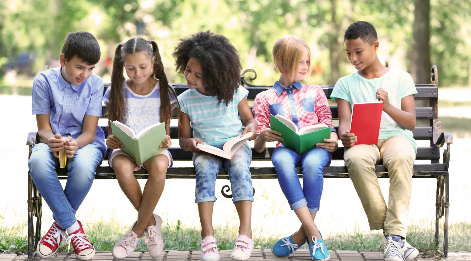 Five children of different races and genders sitting on a park bench reading books.